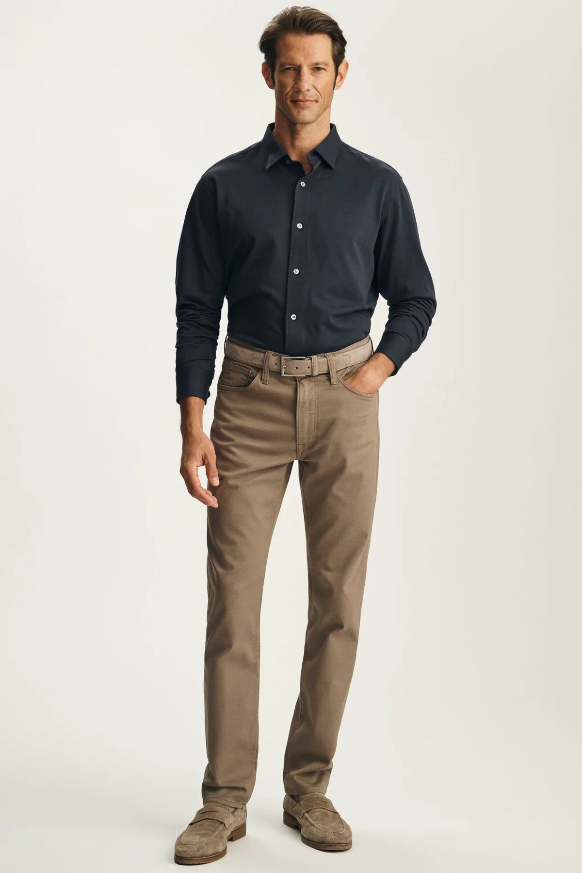 A man modeling a dark navy long-sleeve button-down shirt and tan chino pants with brown shoes, standing against a light beige studio background.