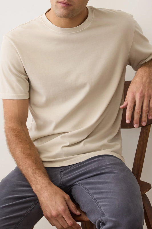 Man wearing a beige t-shirt and gray pants sitting on a wooden chair against a plain background