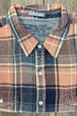 A folded long-sleeve plaid button-up shirt with a collar, laid flat on a gray wood-textured surface. The shirt features a mix of dusty pink, navy blue, and beige plaid patterns with white buttons.
