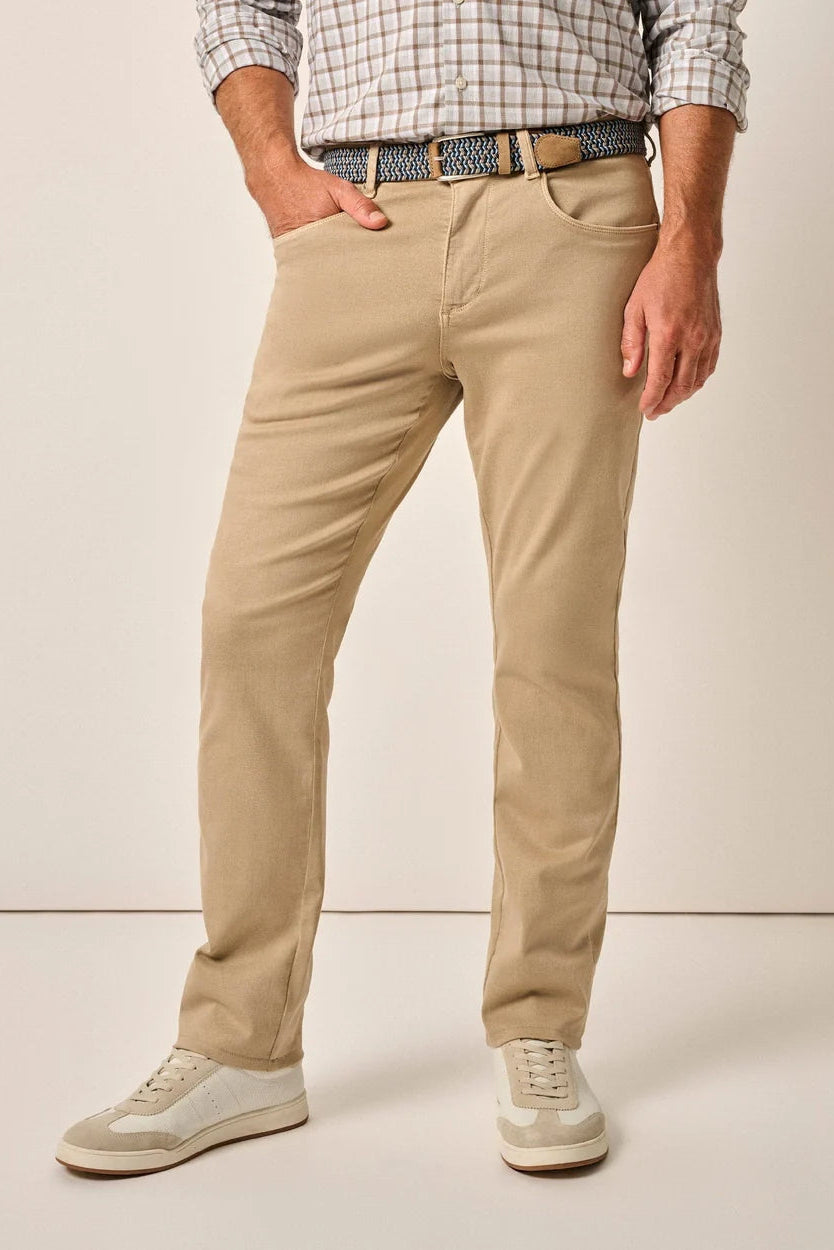 A man wearing beige slim-fit chino pants, a patterned belt, a checkered shirt, and light-colored sneakers, standing against a plain beige studio background.