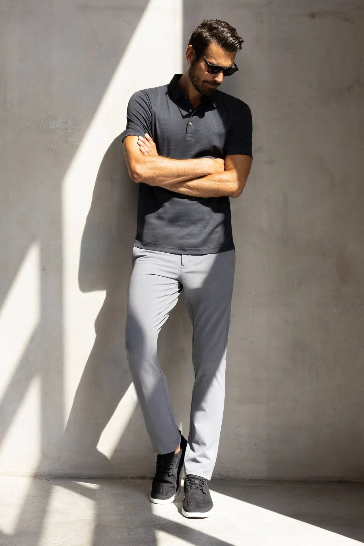 Man wearing a gray polo shirt and light gray pants against a concrete wall.