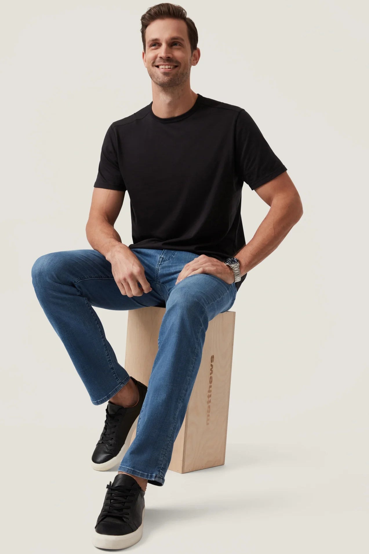 Man sitting on a cardboard box wearing a black t-shirt and blue jeans against a plain background