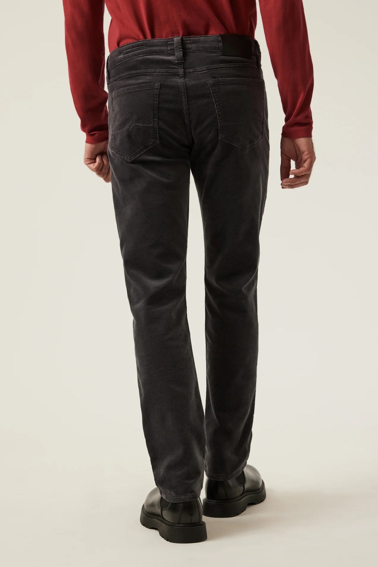 Rear view of a person wearing dark charcoal gray corduroy trousers, a red long-sleeve shirt, and black shoes against a plain light beige studio background.