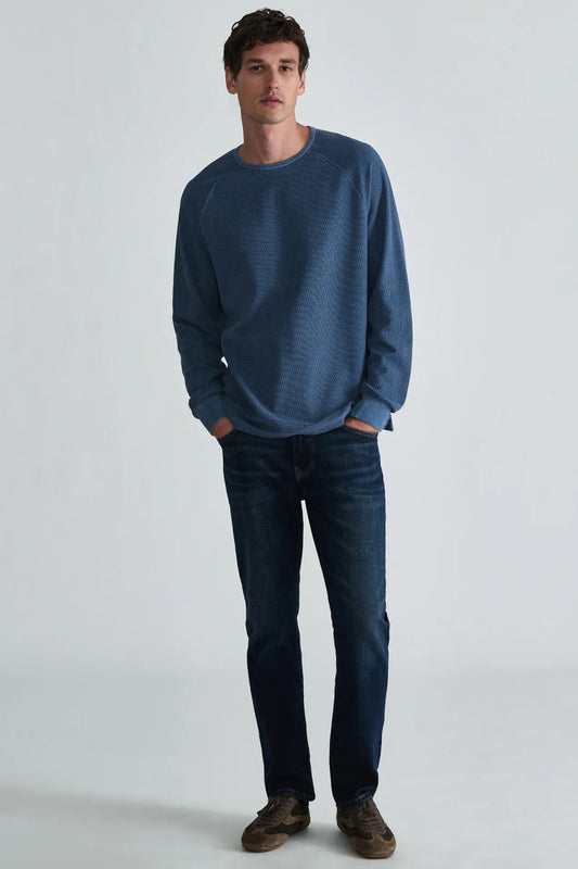 A man wearing a textured blue long-sleeve crewneck sweater, dark blue jeans, and brown shoes, standing against a light gray studio background.