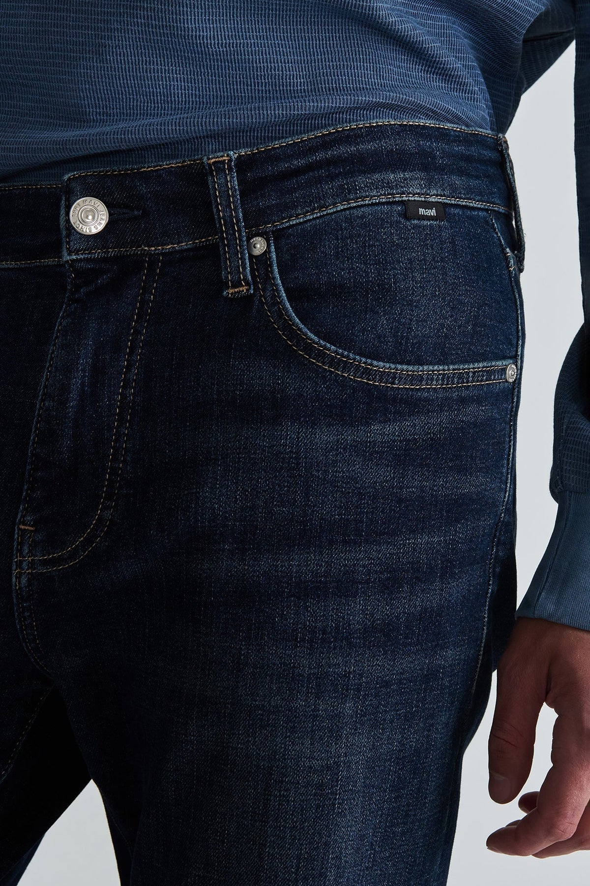 Close-up of a person wearing dark indigo blue denim jeans with a blue textured shirt, showing the waistband, button, belt loops, and front pocket.