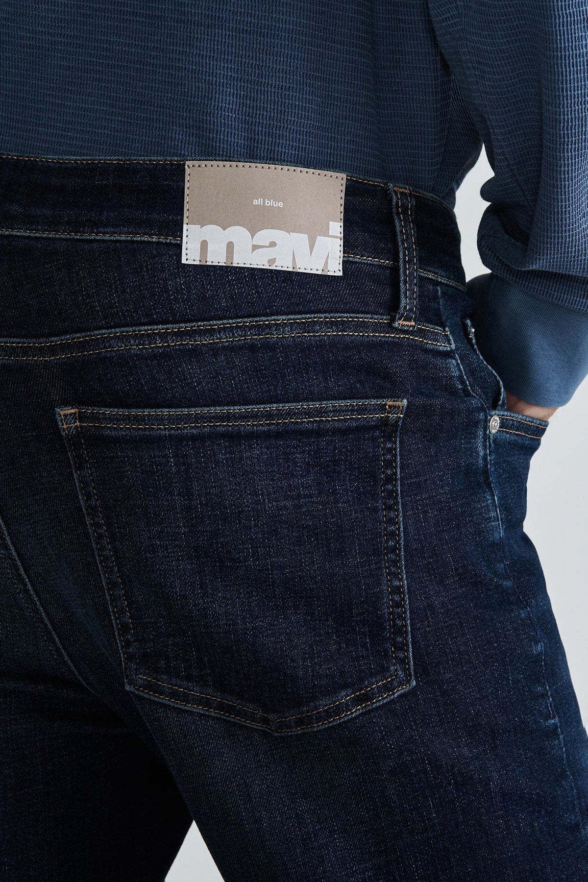 Close-up back view of a person wearing dark indigo denim jeans with a tan Mavi brand patch at the waistband and a blue textured shirt.
