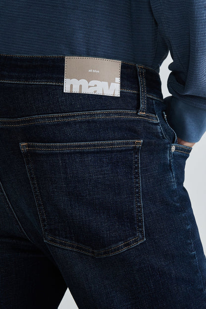 Close-up back view of a person wearing dark indigo denim jeans with a tan Mavi brand patch at the waistband and a blue textured shirt.