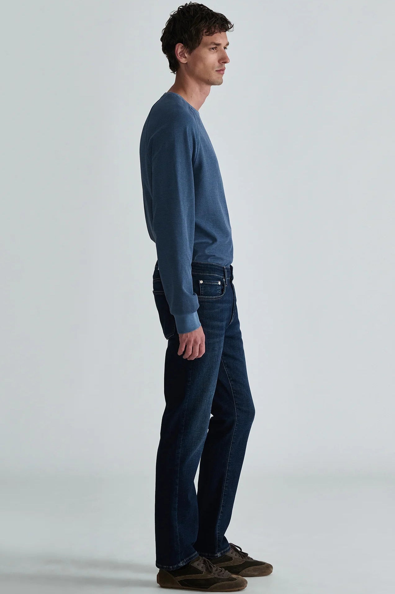 A man in profile wearing a slate blue long-sleeve knit sweater, dark blue jeans, and brown suede shoes, standing against a light gray studio background.