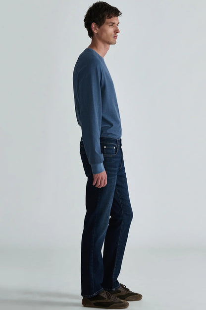 A man in profile wearing a slate blue long-sleeve knit sweater, dark blue jeans, and brown suede shoes, standing against a light gray studio background.