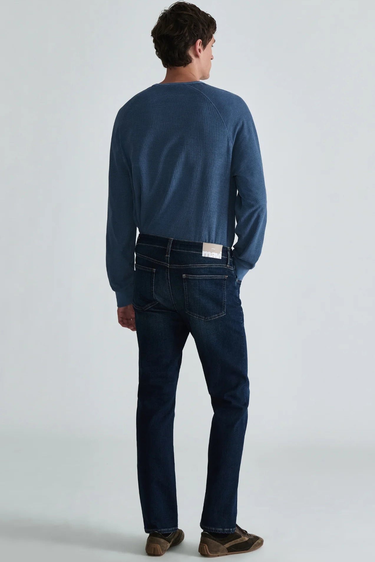 A man viewed from behind wearing a blue long-sleeve knit top, dark blue jeans, and brown shoes, standing against a light gray studio background.