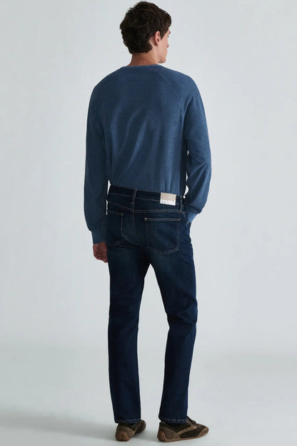 A man viewed from behind wearing a blue long-sleeve knit top, dark blue jeans, and brown shoes, standing against a light gray studio background.