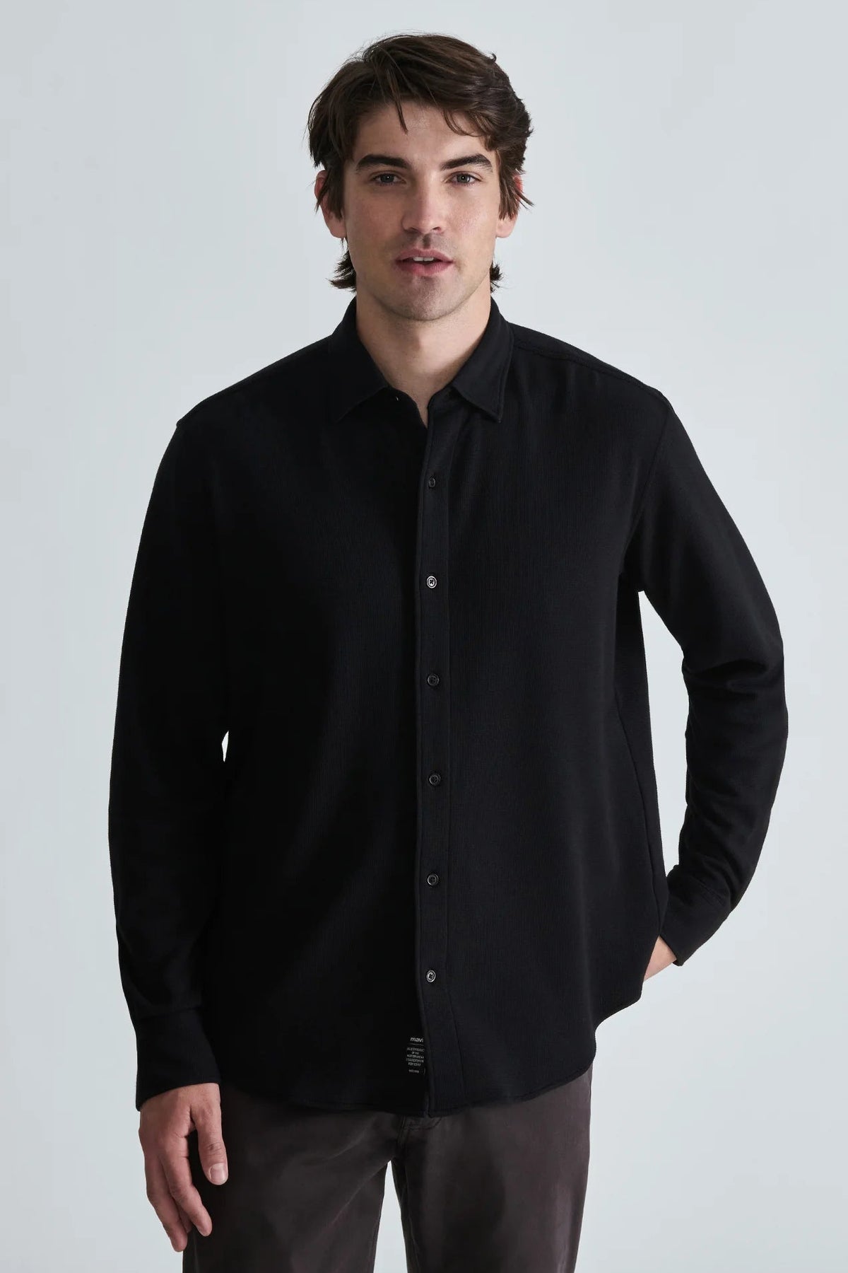 A man wearing a black long-sleeve button-down shirt and dark gray trousers, standing against a light gray studio background.