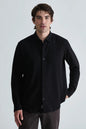 A man wearing a black long-sleeve button-down shirt and dark gray trousers, standing against a light gray studio background.