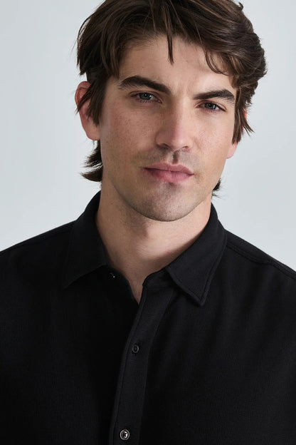 A close-up portrait of a man with brown hair wearing a black button-down shirt against a light gray background.