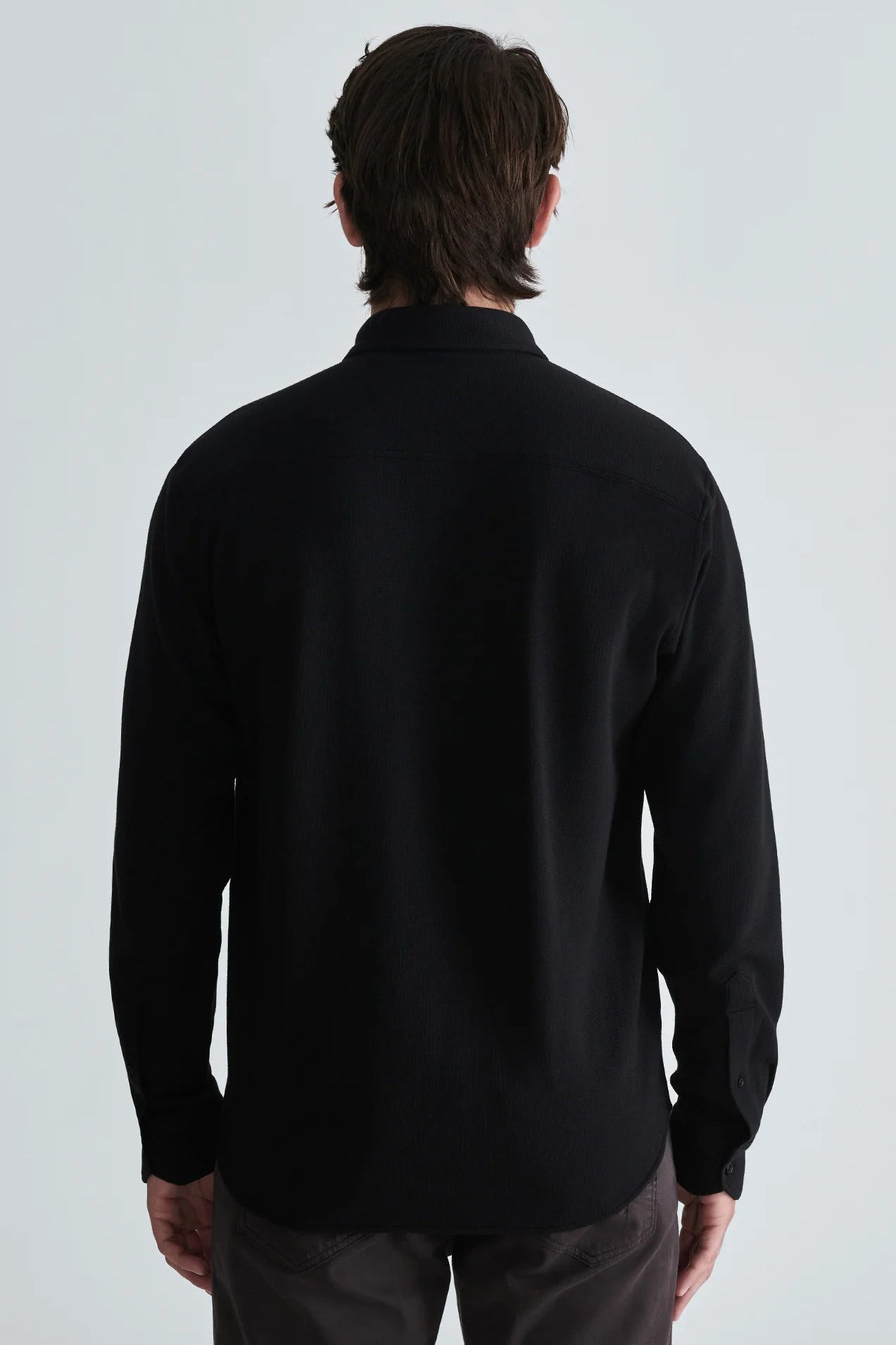 A man viewed from the back wearing a black long-sleeve button-up shirt and dark trousers against a light gray studio background.