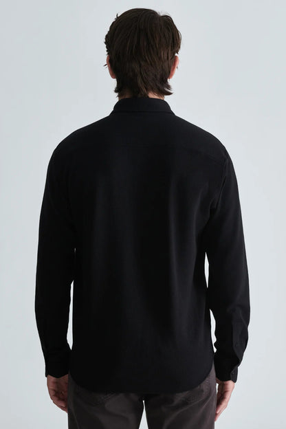 A man viewed from the back wearing a black long-sleeve button-up shirt and dark trousers against a light gray studio background.