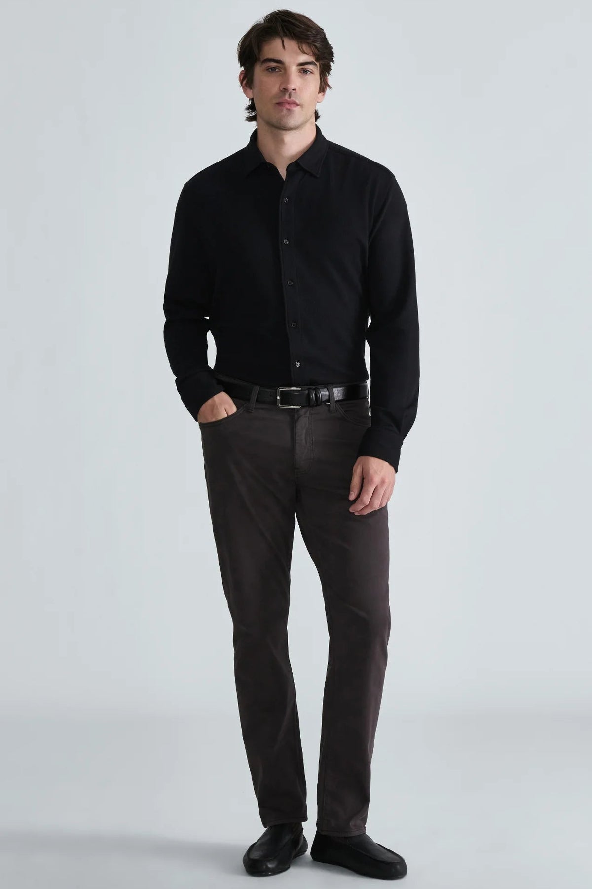A man modeling a black long-sleeve button-down shirt paired with dark brown trousers, a black belt, and black dress shoes, standing against a light gray studio background.