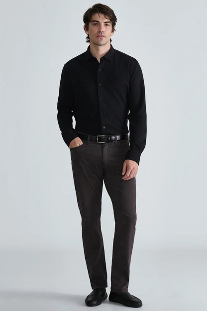 A man modeling a black long-sleeve button-down shirt paired with dark brown trousers, a black belt, and black dress shoes, standing against a light gray studio background.