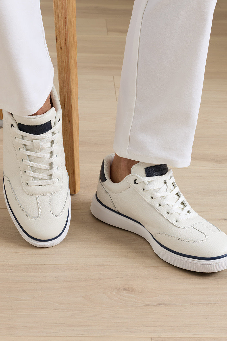 A person wearing off-white leather low-top sneakers with navy blue accents, paired with white trousers, standing on a light wood floor.