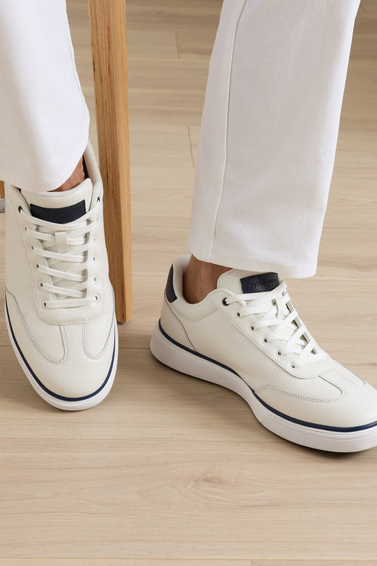 A person wearing off-white leather low-top sneakers with navy blue accents, paired with white trousers, standing on a light wood floor.