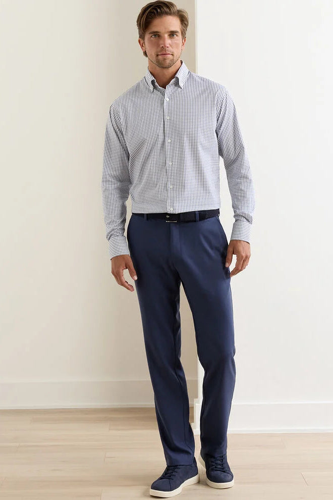 Man wearing a checkered shirt and navy pants standing against a white wall.