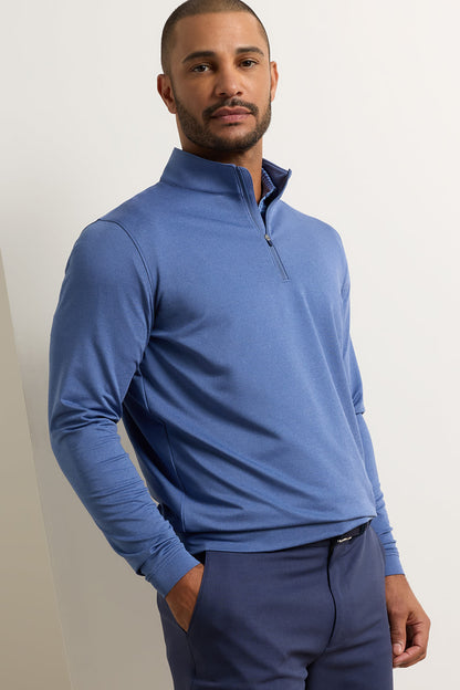 A man wearing a blue long-sleeve quarter-zip pullover and matching blue trousers, standing against a plain off-white wall.