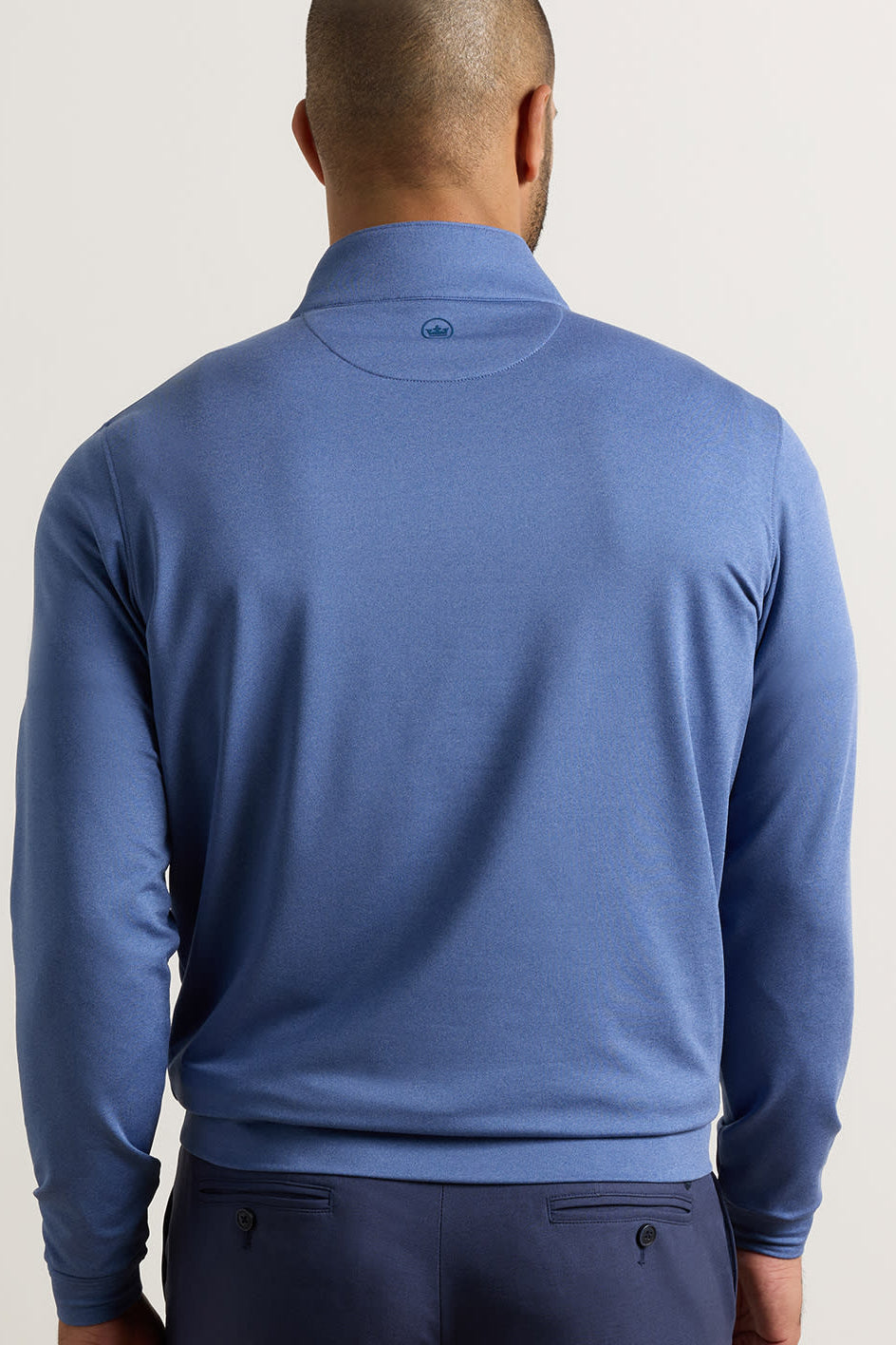 A man viewed from the back wearing a blue long-sleeve quarter-zip pullover and navy pants, against a plain light background.