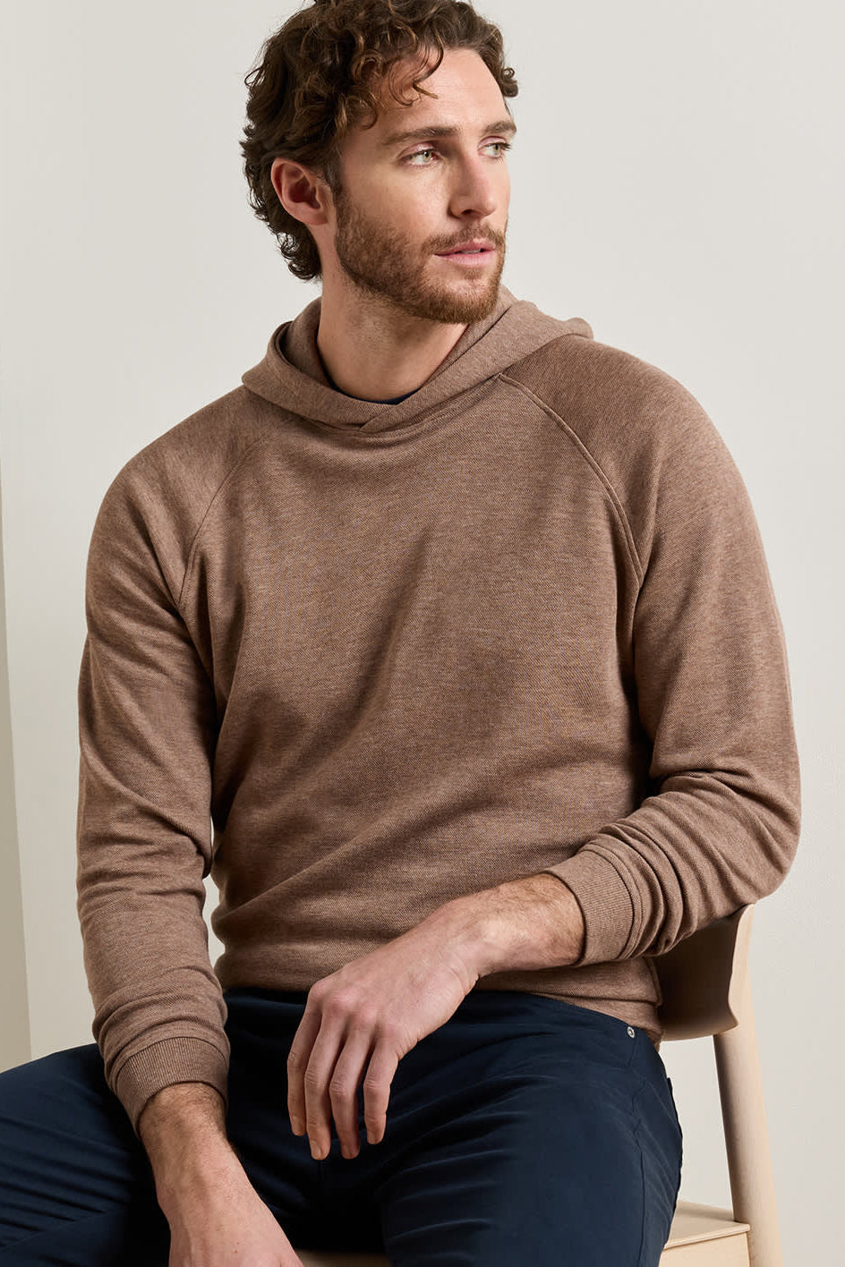 A man wearing a brown long-sleeve knit hoodie and navy pants, sitting on a light-colored wooden chair against a neutral beige background.