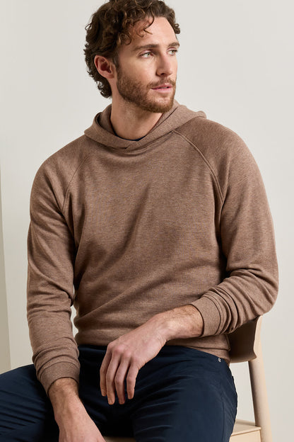 A man wearing a brown long-sleeve knit hoodie and navy pants, sitting on a light-colored wooden chair against a neutral beige background.
