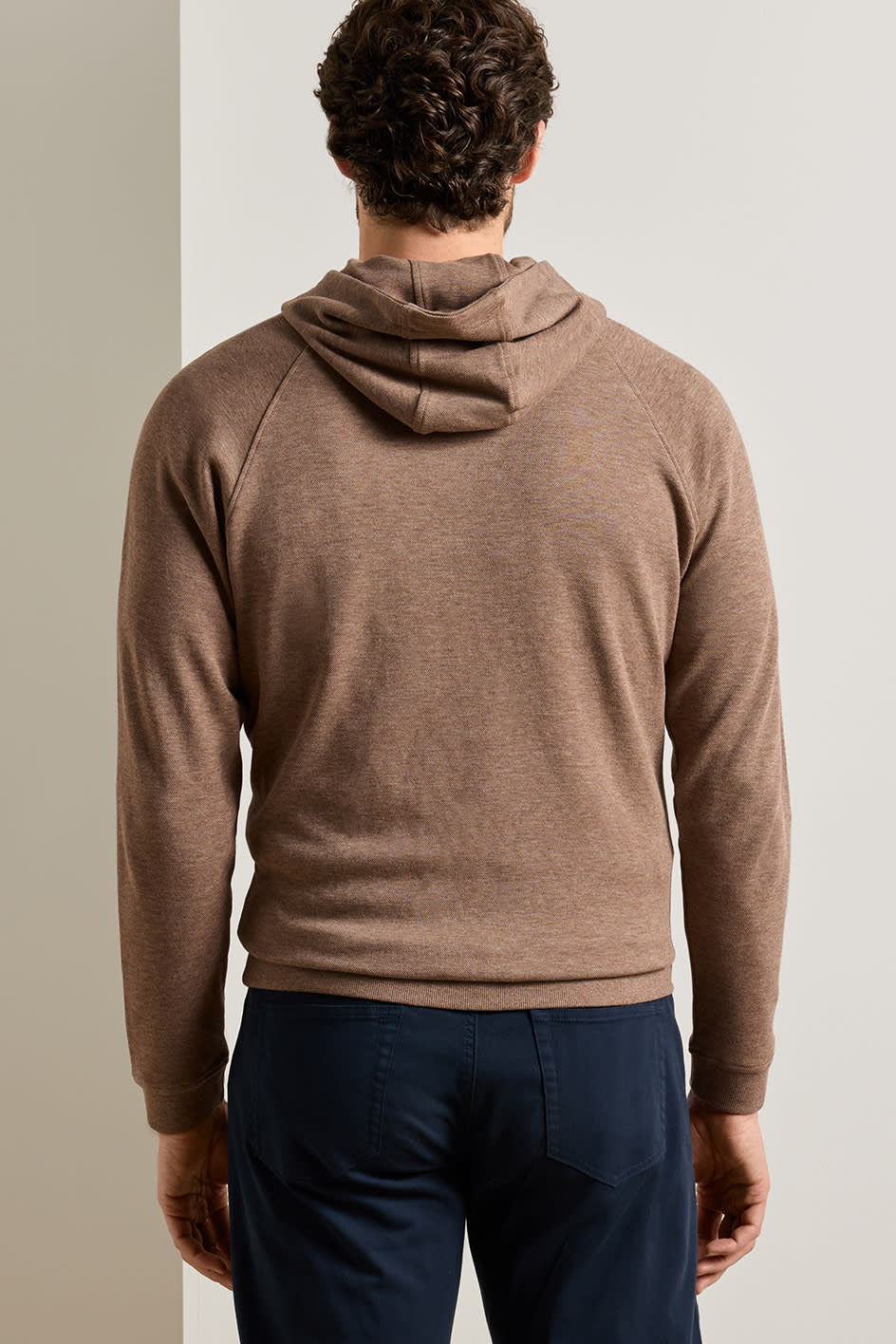 A man viewed from behind wearing a brown knit hooded sweater and navy blue trousers, standing against a neutral light gray background.