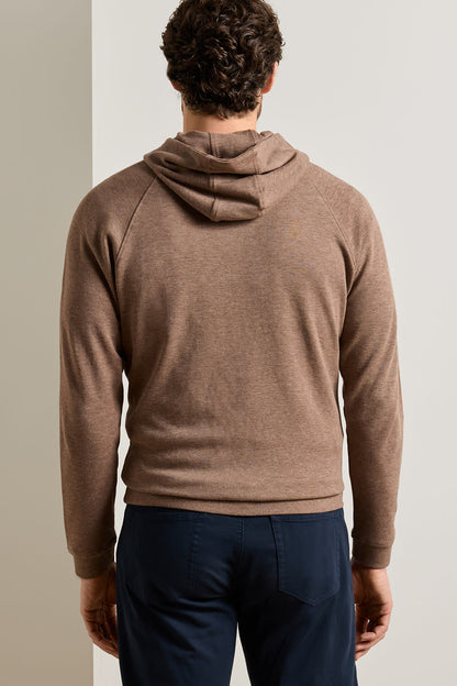 A man viewed from behind wearing a brown knit hooded sweater and navy blue trousers, standing against a neutral light gray background.
