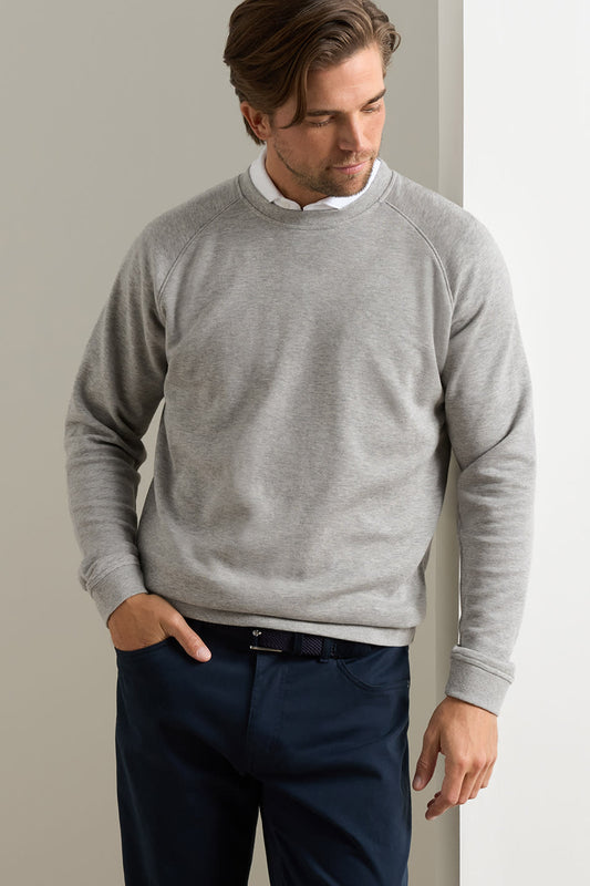 A man wearing a light gray crewneck sweater over a white collared shirt and navy blue trousers, standing against a neutral light gray wall.