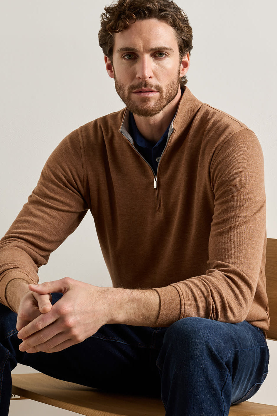 A man sitting on a wooden chair wearing a brown quarter-zip long-sleeve sweater over a navy shirt and blue jeans, photographed against a plain light gray studio background.
