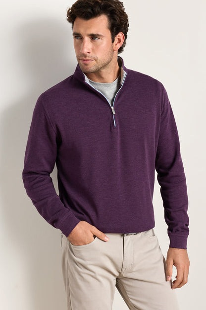 A man wearing a dark purple quarter-zip long-sleeve sweater over a light gray t-shirt and beige pants, standing against a plain off-white wall.