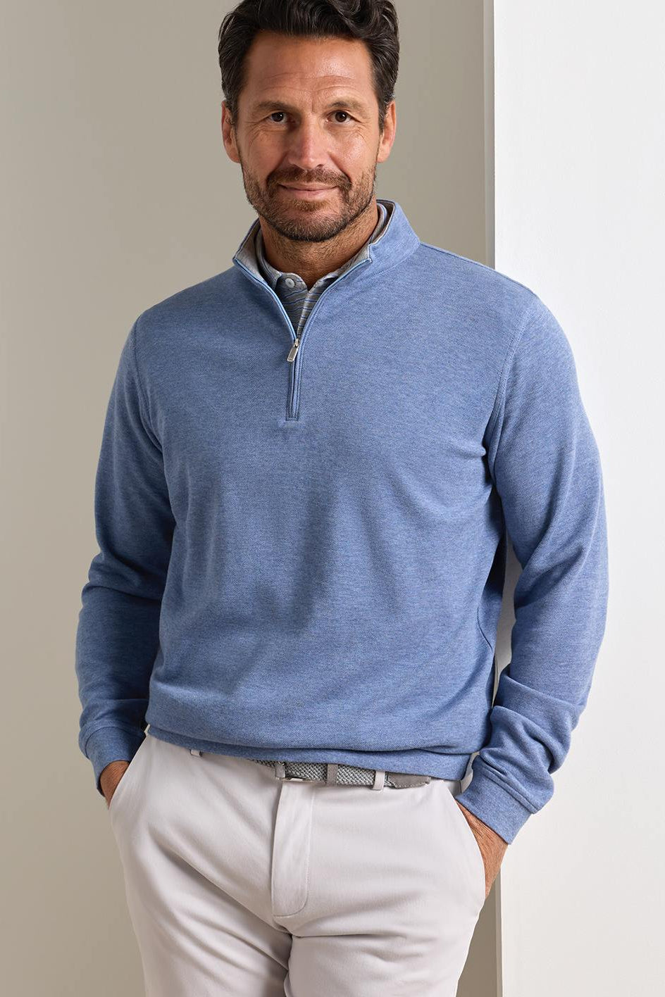A man wearing a heathered blue quarter-zip long-sleeve pullover sweater and white trousers, standing against a neutral light gray background.