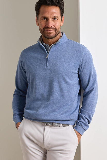 A man wearing a heathered blue quarter-zip long-sleeve pullover sweater and white trousers, standing against a neutral light gray background.