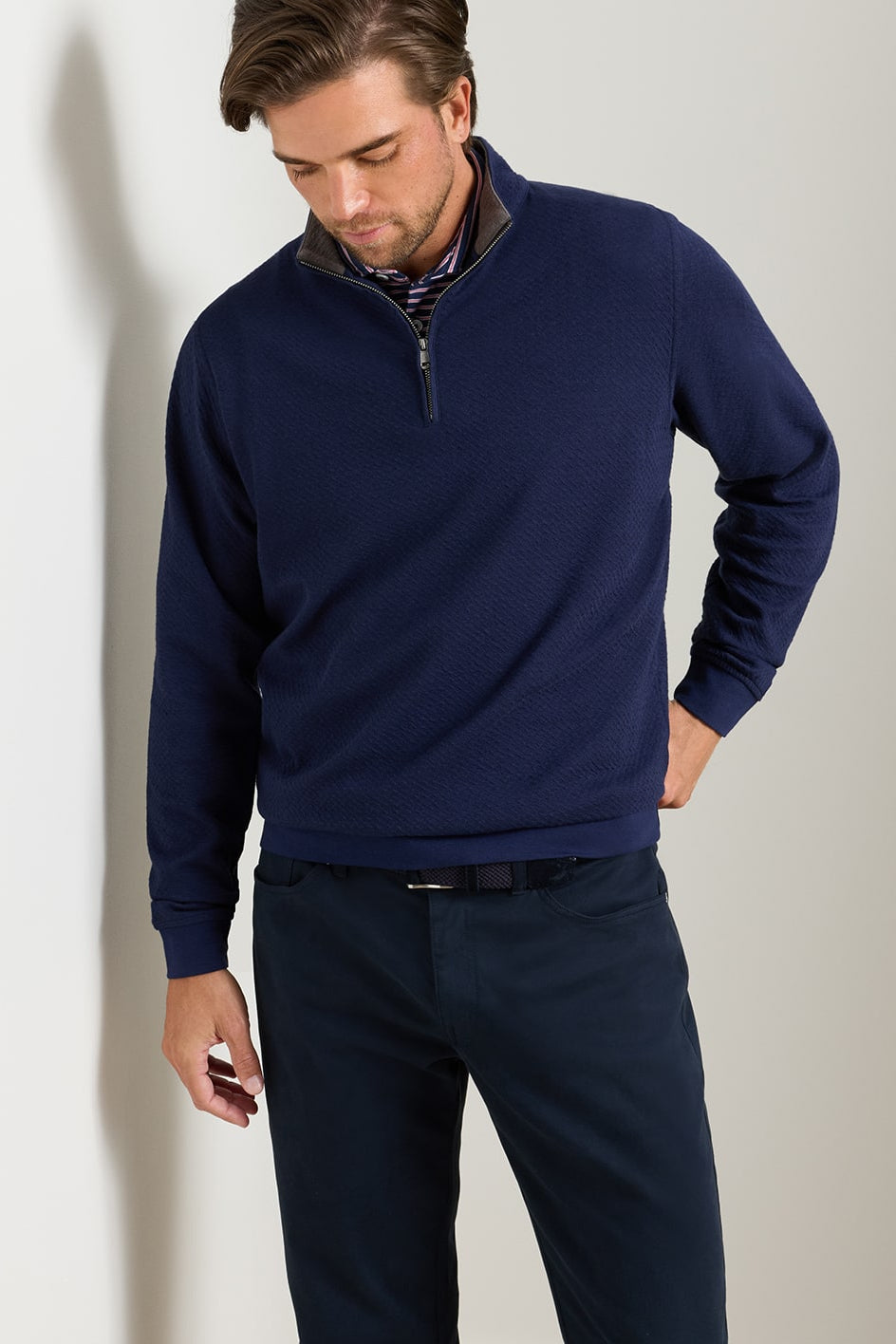 A man wearing a navy blue quarter-zip pullover sweater with a subtle textured collar, paired with dark trousers, standing against a light gray wall.