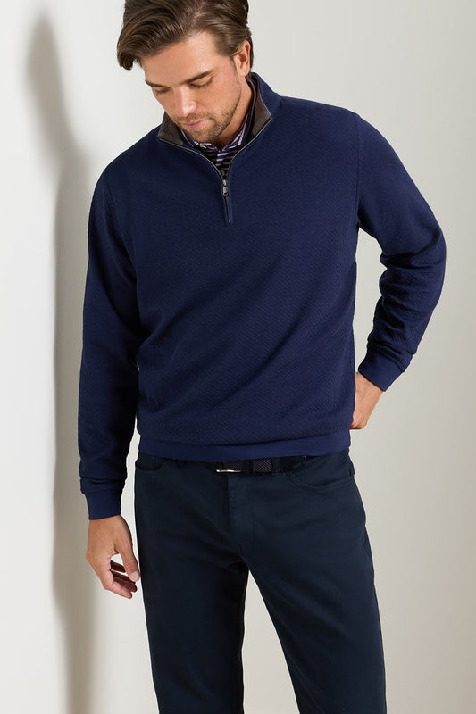 A man wearing a navy blue quarter-zip pullover sweater with a subtle textured collar, paired with dark trousers, standing against a light gray wall.