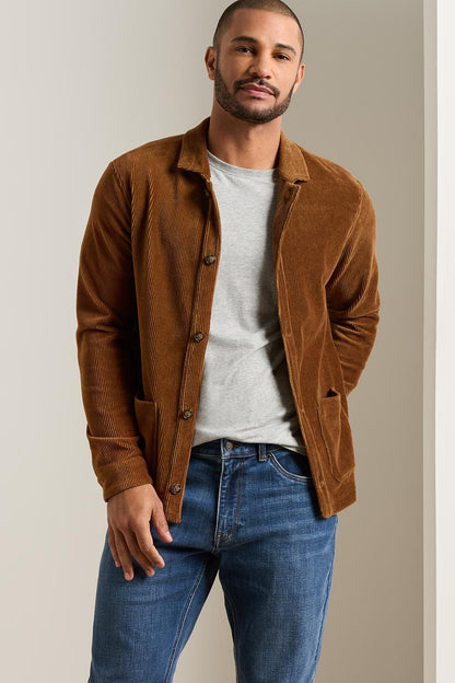 A man wearing a brown corduroy jacket over a light gray t-shirt and blue jeans, standing against a neutral beige background.