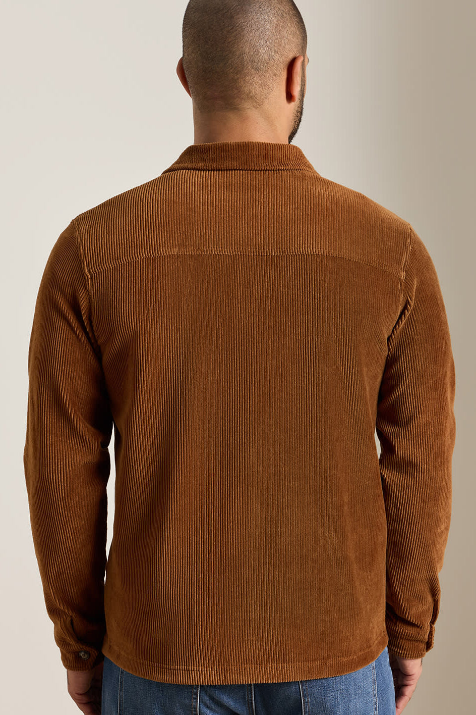 A man viewed from the back wearing a brown corduroy shirt jacket, paired with blue jeans, against a plain beige studio background.