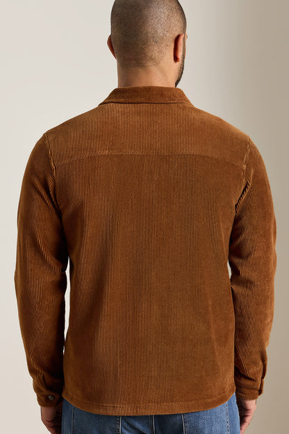 A man viewed from the back wearing a brown corduroy shirt jacket, paired with blue jeans, against a plain beige studio background.