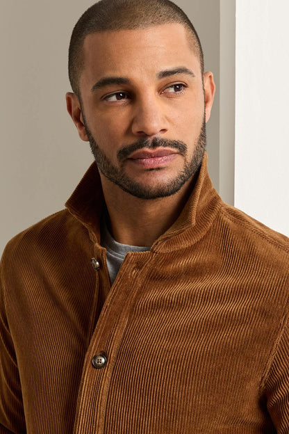 A man wearing a brown corduroy button-front jacket over a gray t-shirt, photographed in a clean indoor setting with a light gray wall.