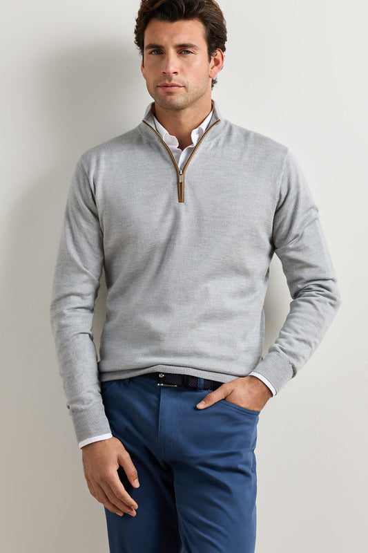 A man wearing a light gray quarter-zip sweater over a white collared shirt and blue trousers, standing against a plain off-white wall.