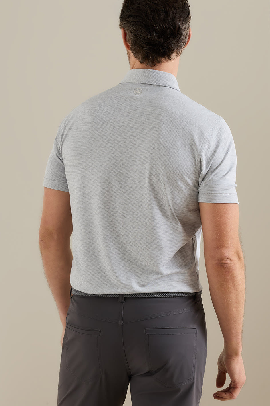 A man viewed from behind wearing a light gray short-sleeve polo shirt and dark gray trousers, standing against a plain beige wall.