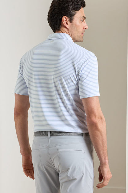 A man shown from the back wearing a light blue and white striped short-sleeve polo shirt with light gray trousers, standing against a neutral beige wall.