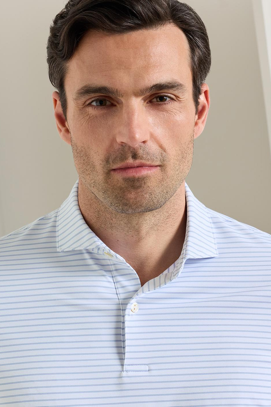 A close-up shot of a man wearing a light blue and white striped polo shirt, photographed against a soft beige background.