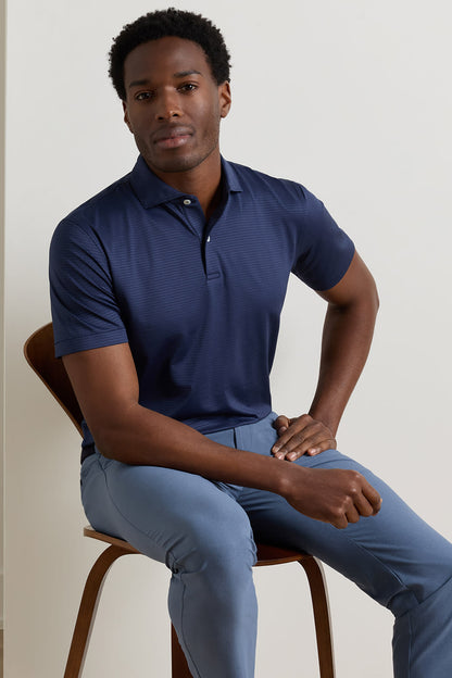 A man wearing a navy blue short-sleeve polo shirt and light blue trousers, seated on a wooden chair against a plain off-white wall.