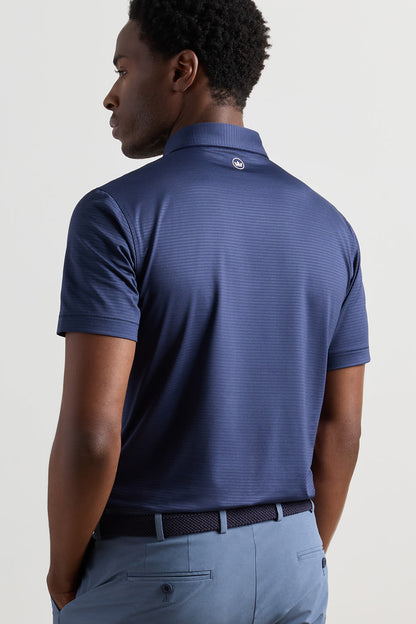 A man shown from the back wearing a navy blue short-sleeve polo shirt with a subtle textured pattern, paired with light blue trousers, against a plain light gray studio background.