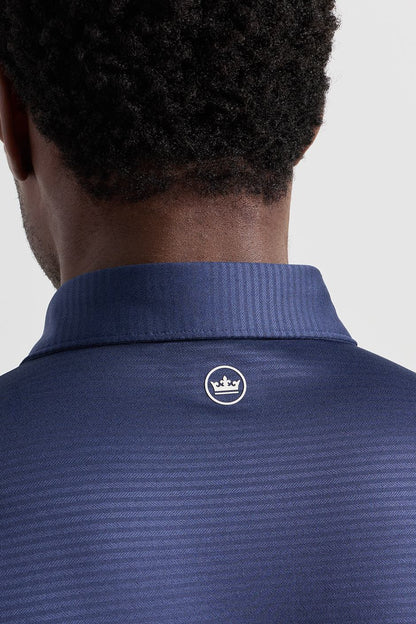 Close-up back view of a man wearing a textured navy blue polo shirt with a ribbed collar and a small white crown logo on the upper back.