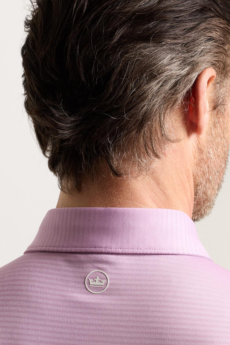 Close-up back view of a man wearing a light pink polo shirt, showing the textured fabric, collar, and a small circular logo on the upper back.